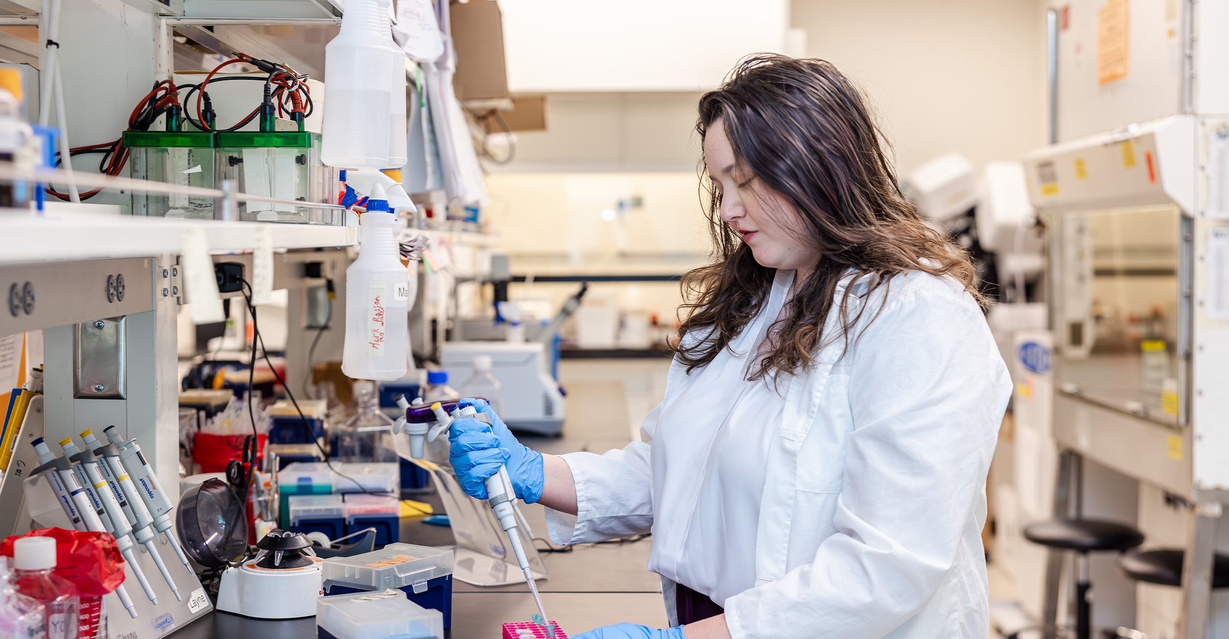 meeker_banner A laboratory technician wearing a white lab coat and blue gloves uses a pipette to transfer liquid into a sample tray at a well‑equipped research bench inside a laboratory.