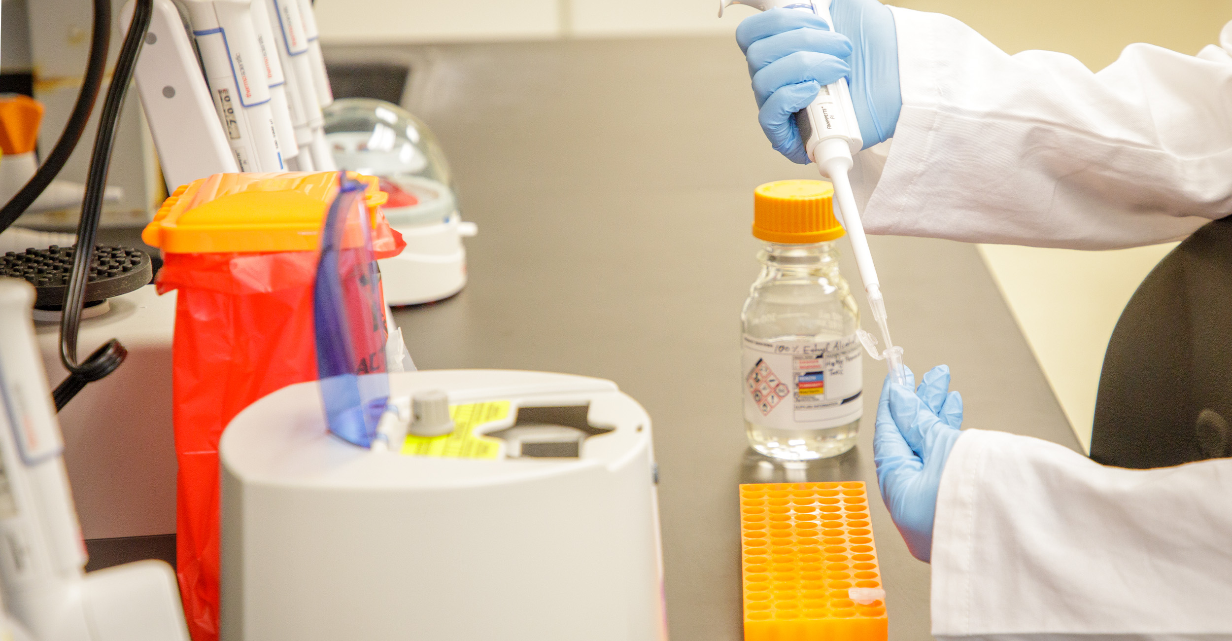 ngscf_banner Gloved hands use a pipette to transfer liquid into a small tube at a laboratory bench organized with vials, racks, and scientific equipment, illustrating a precise lab procedure.