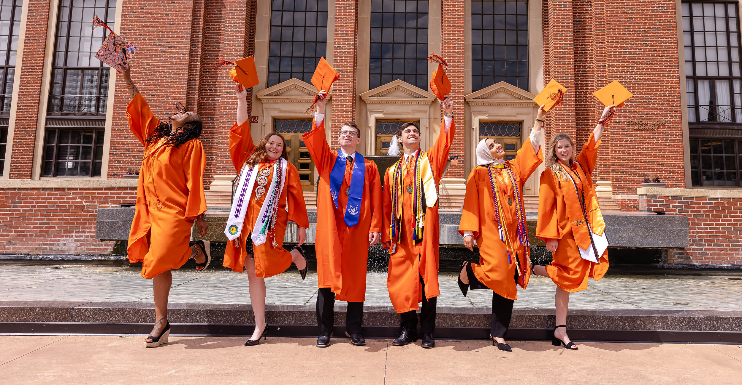 orange_gown_banner Six graduates in orange caps and gowns stand in front of a brick campus building and fountain, holding their caps in the air in a celebratory pose during a graduation photo.