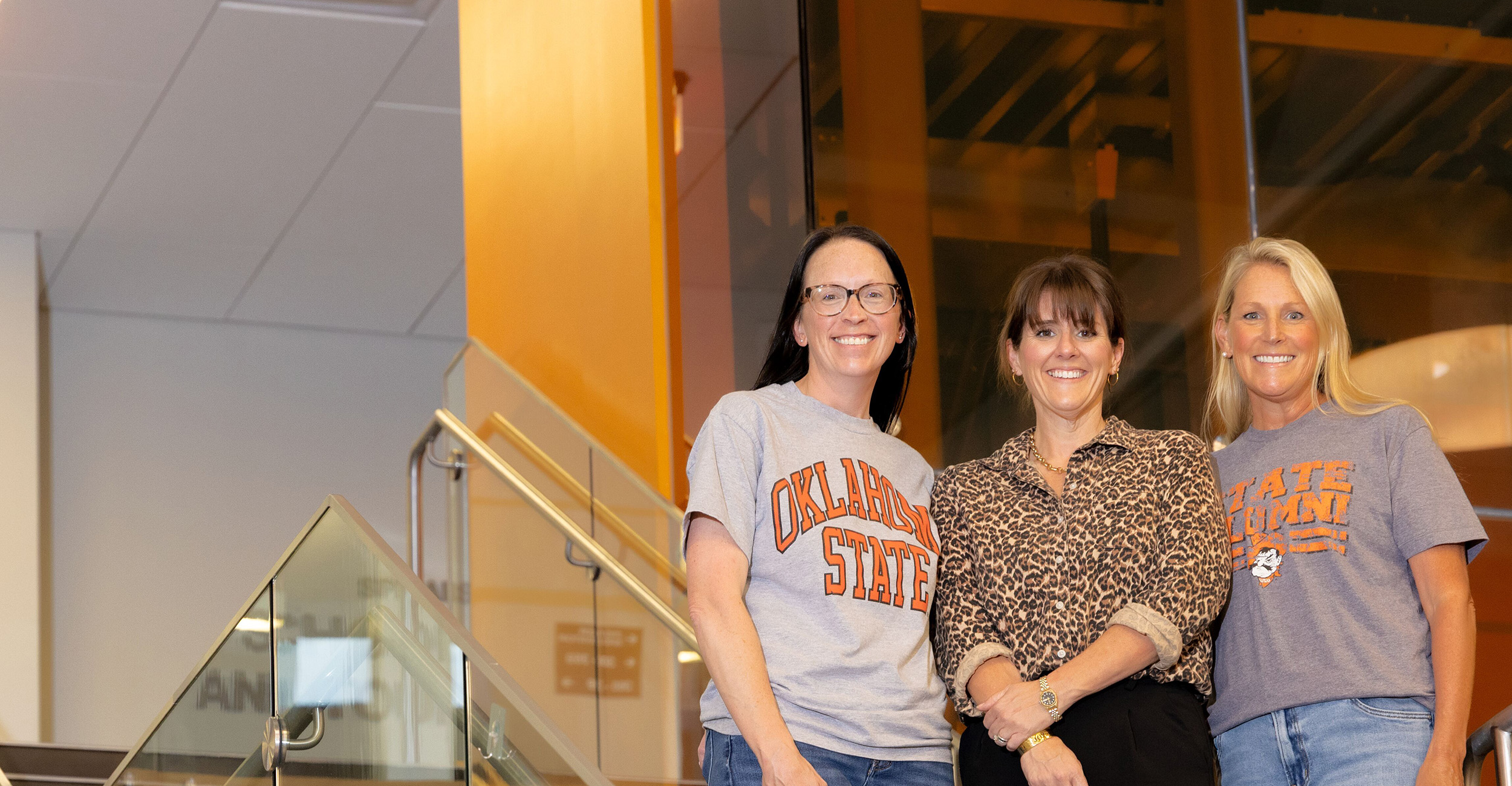 principals_banner_3 Three people stand together on an indoor staircase with glass railings, wearing Oklahoma State University–branded shirts in a modern building interior.