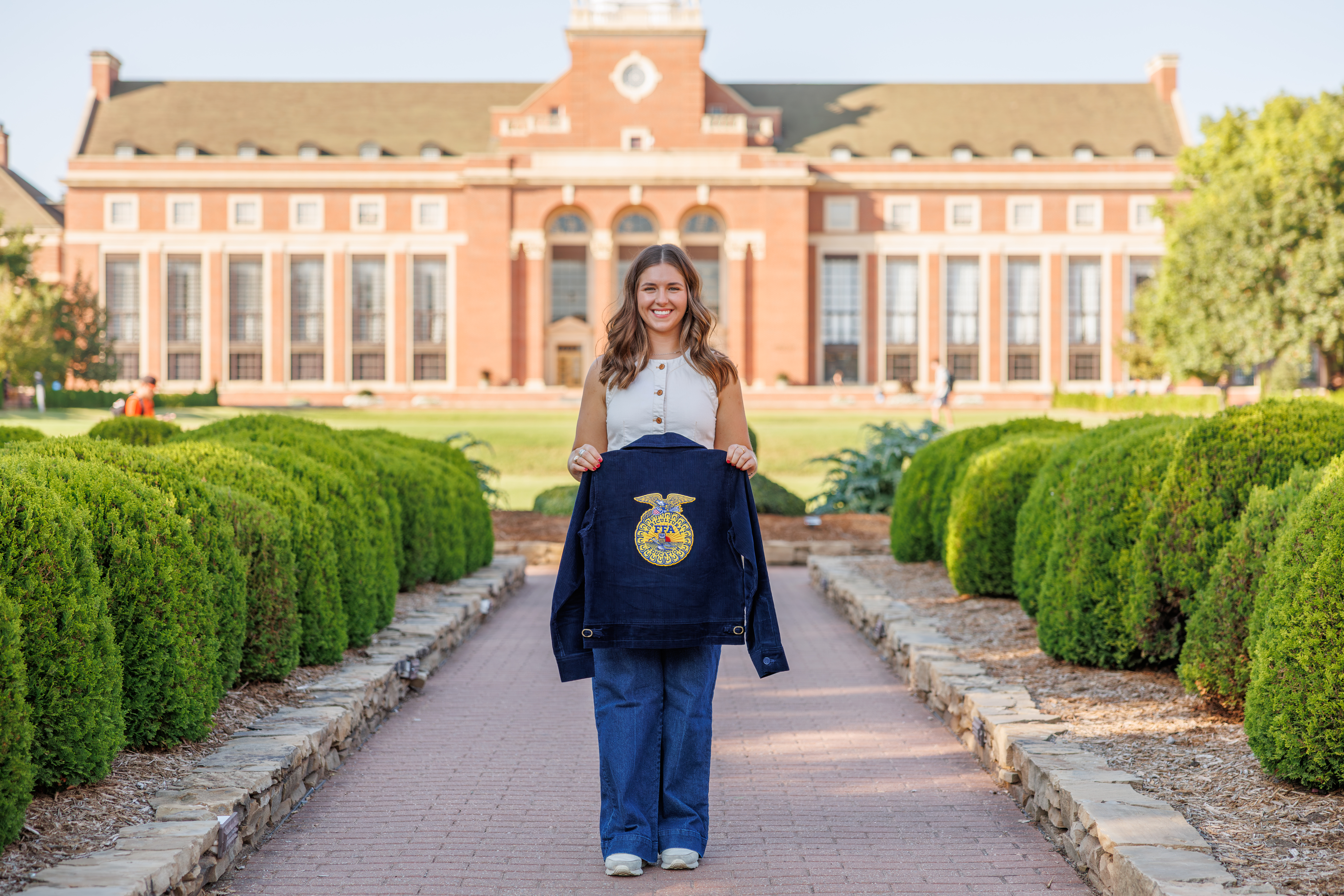 A person stands centered on a campus walkway in front of a large academic building, holding a blue FFA jacket toward the camera, with neatly trimmed bushes lining the path and the building’s columns and windows visible in the background.