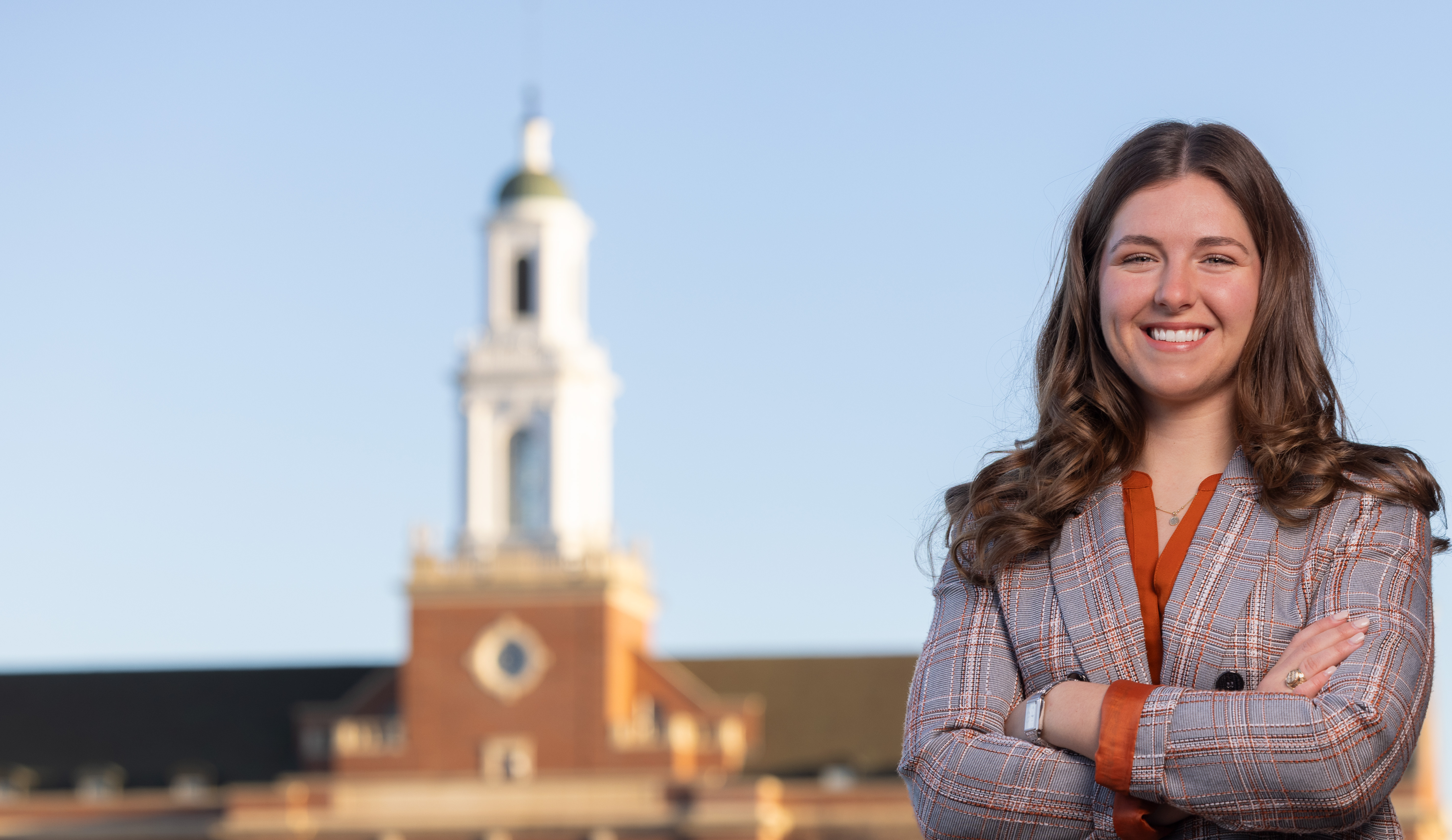 A person in a plaid blazer and orange shirt stands with arms crossed, posed for a professional portrait in front of a blurred campus building with a prominent tower in the background.