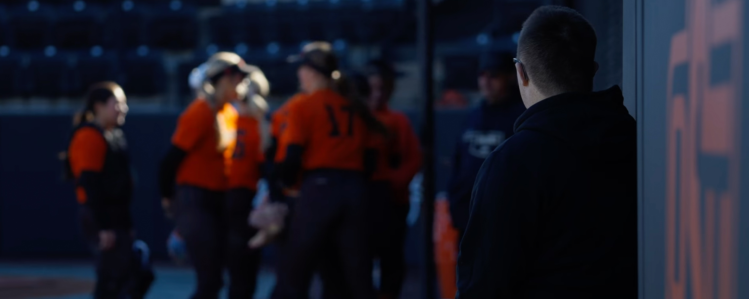 A person stands in the foreground watching a group of players in orange jerseys gathered on a field in the background.