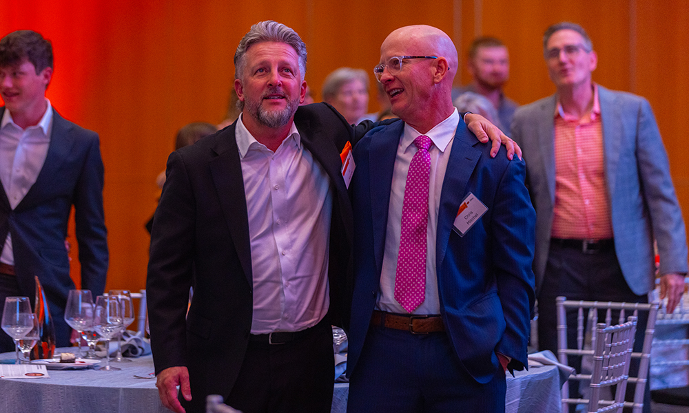 Two men in suits stand arm in arm at a formal banquet or alumni event, with round tables, place settings, and other attendees visible in a warmly lit reception space.