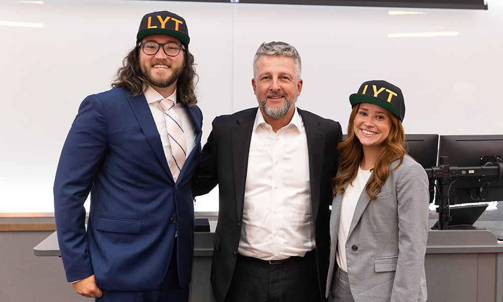 Three professionally dressed individuals stand together in a classroom or lecture space, with two wearing LYT‑branded caps, suggesting collaboration or recognition connected to a business or academic program.