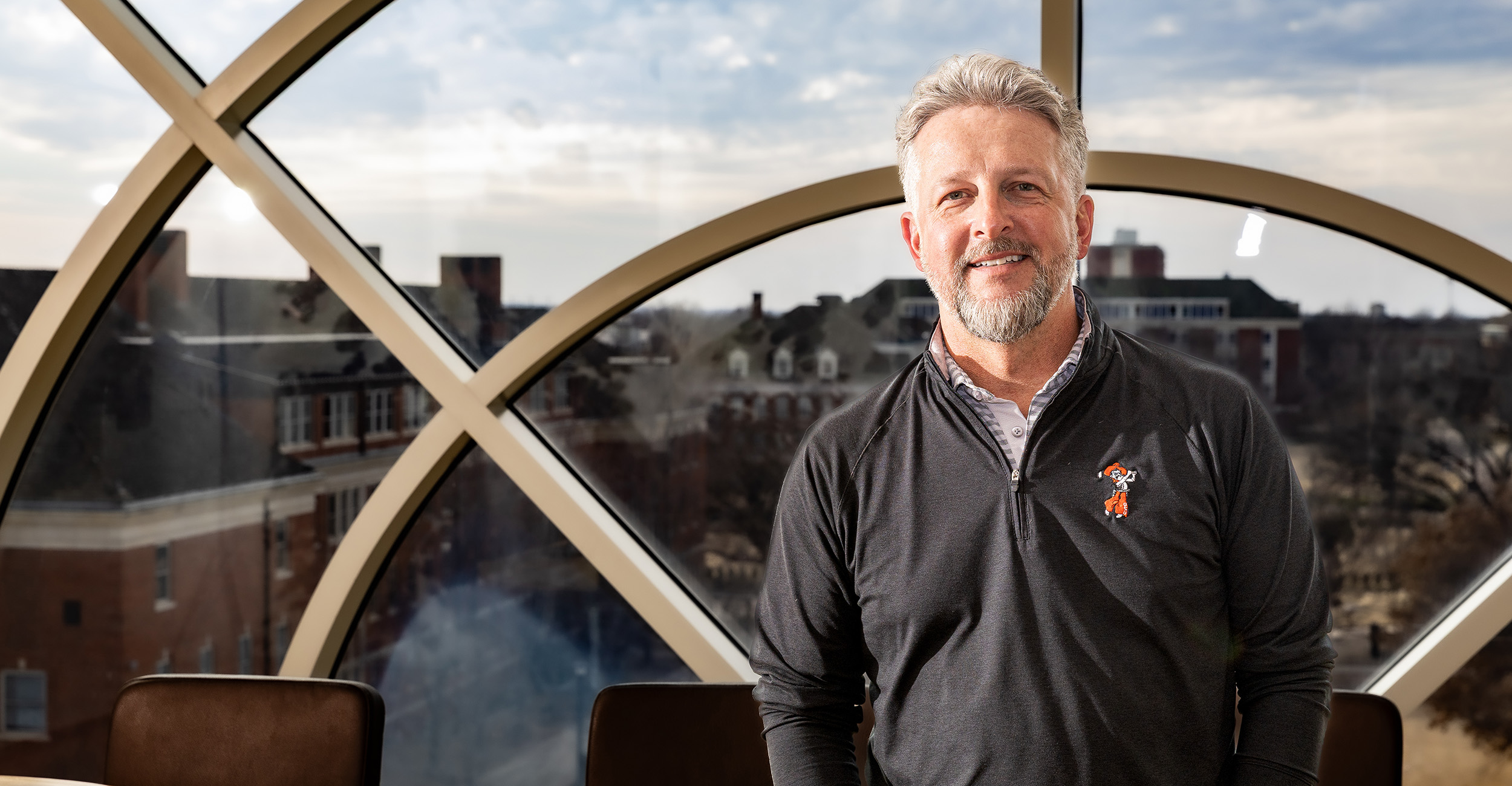 Person wearing a dark quarter‑zip pullover stands indoors in front of a large arched window, with a university campus visible outside, conveying a professional portrait in an academic setting.