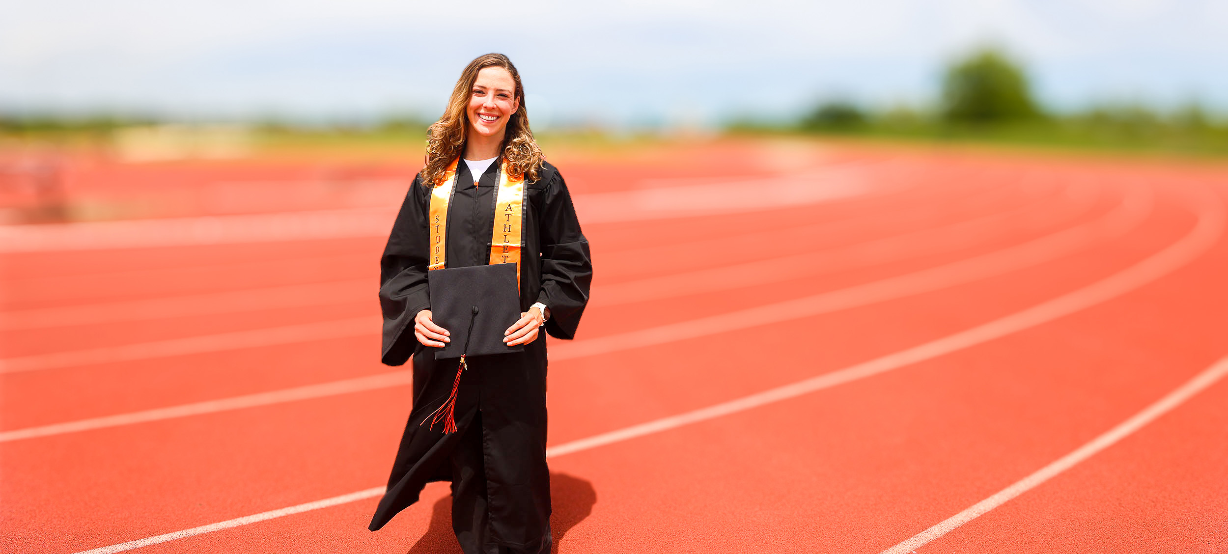 A woman stands on a running track wearing graduation regalia while smiling outdoors. A woman stands on a running track wearing graduation regalia while smiling outdoors.