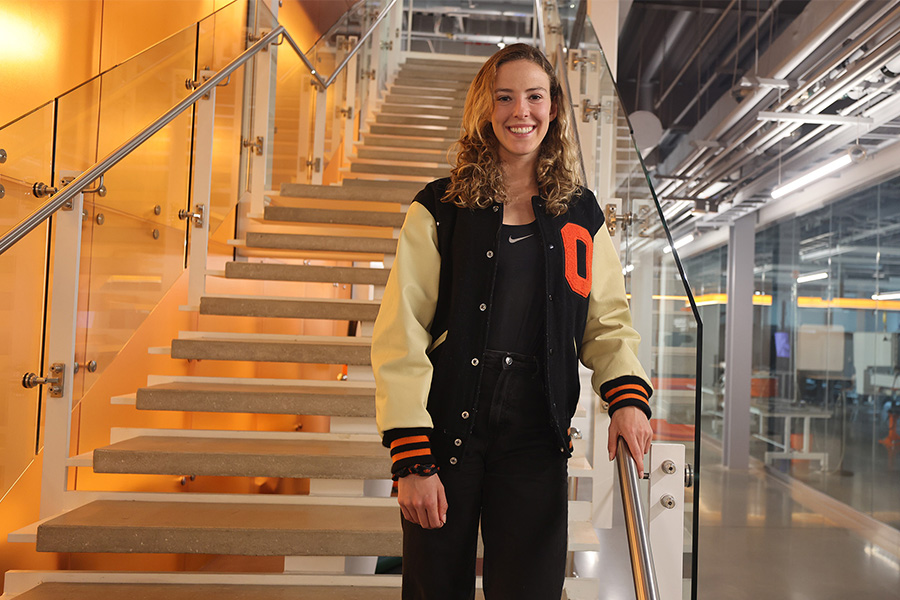 A woman wearing a letterman jacket poses for a photo on a staircase. A woman wearing a letterman jacket poses for a photo on a staircase.