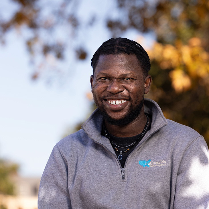 Person wearing a light gray quarter-zip sweatshirt with a blue “Nationwide Marketing Group” logo, layered necklaces, and sitting outdoors with autumn-colored trees in the background.