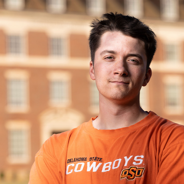 Person wearing an orange Oklahoma State Cowboys T-shirt with OSU logo, standing outdoors in front of a brick building with large windows and architectural details.