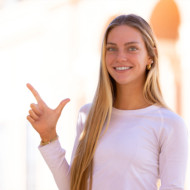 Person with long straight hair wearing a light-colored long-sleeve top and gold earrings, standing outdoors in front of a brick building with arches, making a hand gesture with index finger and thumb extended.