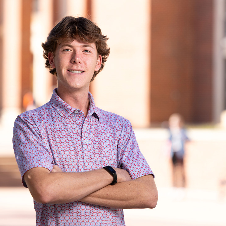 Person wearing a short-sleeve button-up shirt with a small patterned design, standing outdoors in front of a brick building with arms crossed and a smartwatch on the wrist.