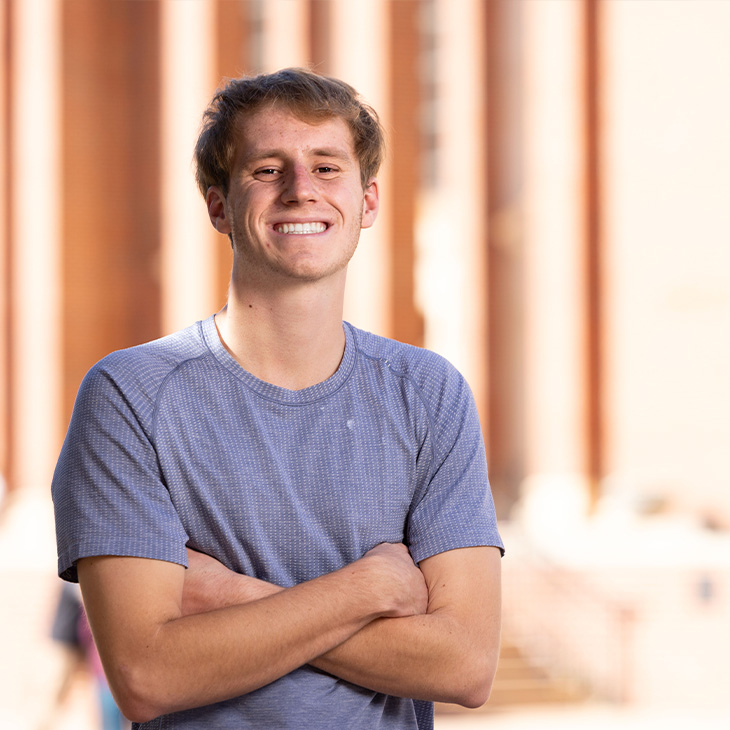 Person wearing a short-sleeve gray textured T-shirt, standing outdoors with arms crossed in front of a brick building with tall columns.