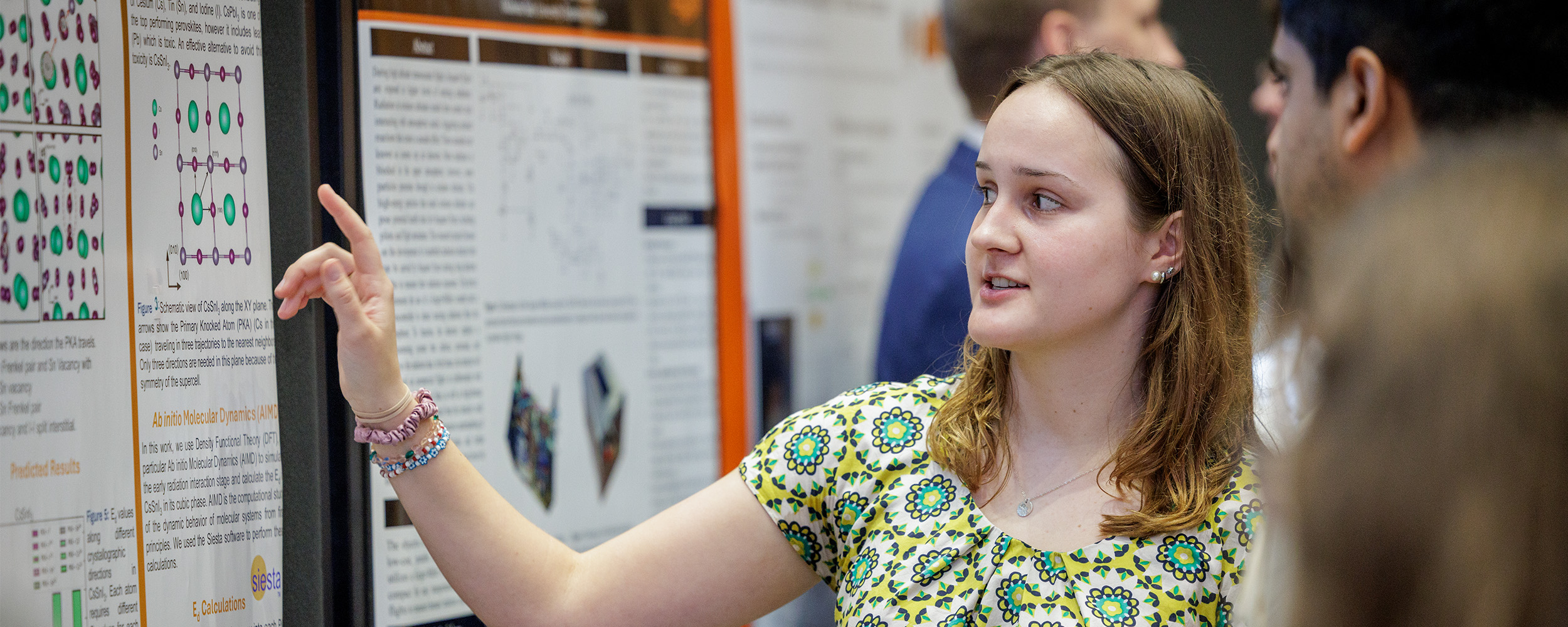  A young woman presenting her research at a symposium, pointing to a colorful academic poster filled with graphs and text. She is wearing a patterned green and yellow shirt and speaking to a small group of people gathered around her. Other research posters and attendees are visible in the background.