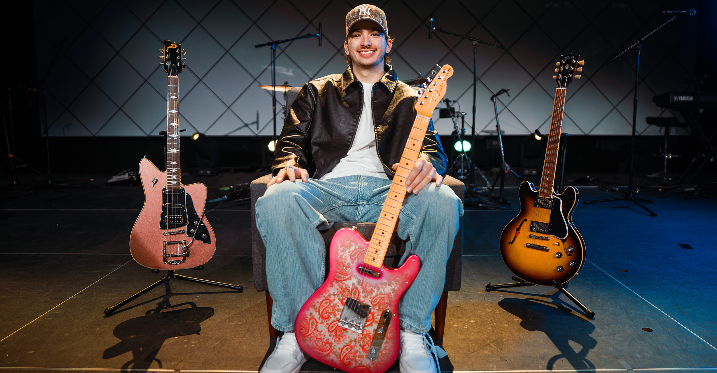  A young man poses on a stage with three guitars. He hold a pink paisley electric guitar, and he also has a guitar on each side of him. Microphones are in the background.