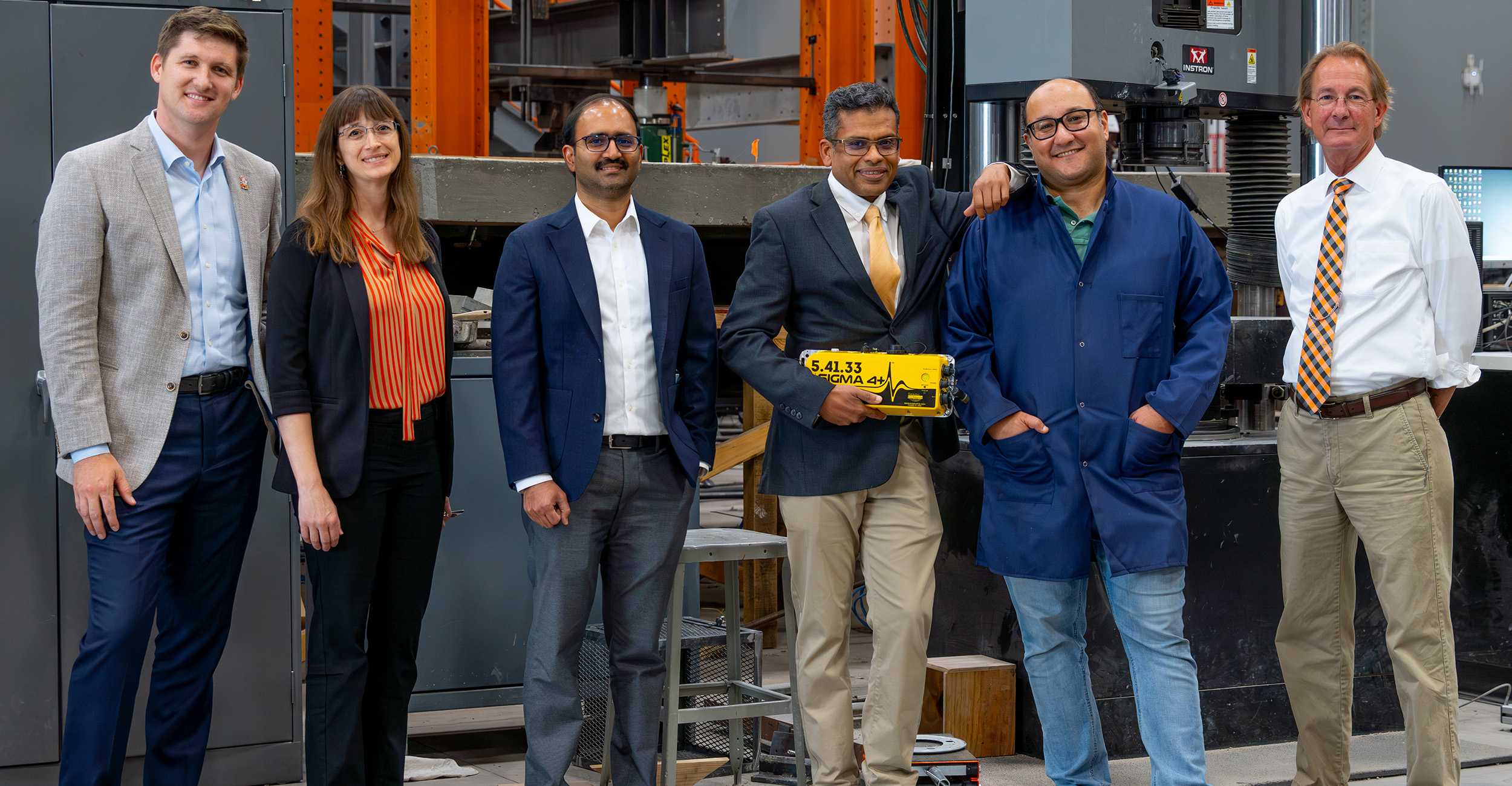 Six individuals standing in a row inside an industrial or laboratory setting, dressed in business casual and professional attire.