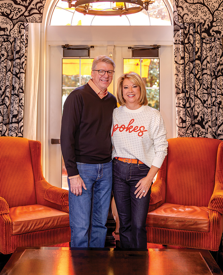 President Jim Hess and his wife Angela pose together in a decorated room with orange upholstered chairs, black and white patterned wallpaper, and large windows. Jim wears a dark V-neck sweater over an orange shirt with jeans, while Angela wears a white sweater with 'Pokes' written in orange letters and dark jeans with an orange belt. President Jim Hess and his wife Angela pose together in a decorated room with orange upholstered chairs, black and white patterned wallpaper, and large windows. Jim wears a dark V-neck sweater over an orange shirt with jeans, while Angela wears a white sweater with 'Pokes' written in orange letters and dark jeans with an orange belt.
