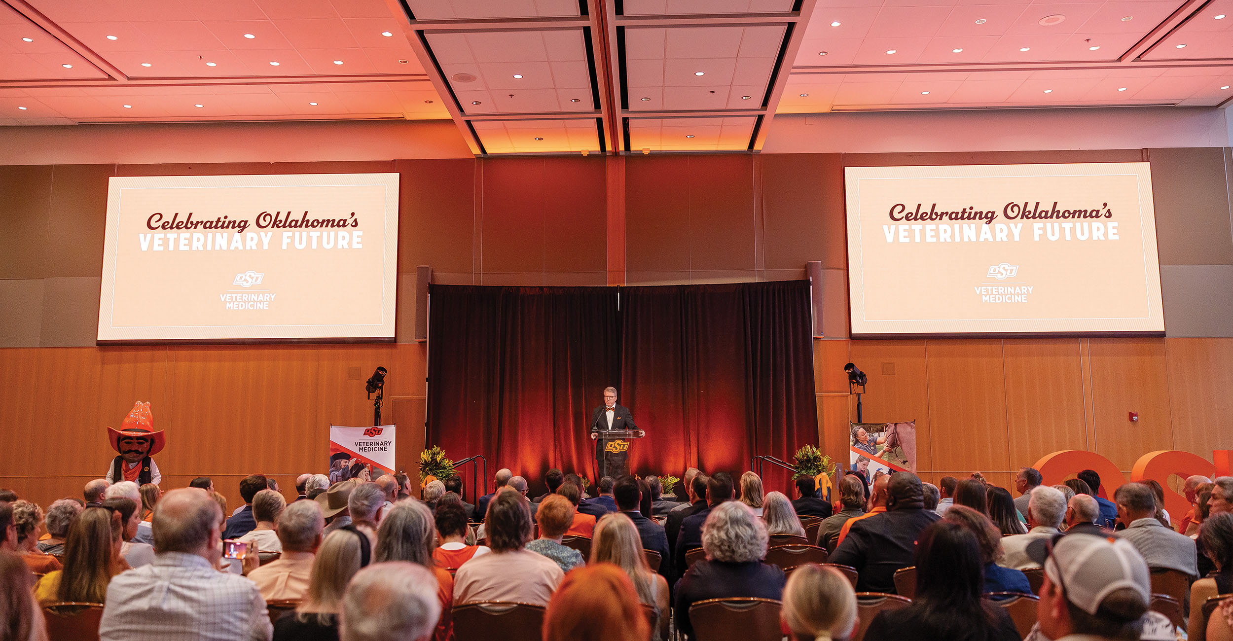  A large audience seated in a conference room faces a stage where a speaker stands at a podium. The stage backdrop features orange lighting and two large screens displaying 'Celebrating Oklahoma's Veterinary Future' with the Oklahoma State University logo and 'Veterinary Medicine.'