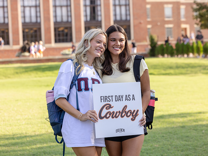 Two ladies stand on a grassy area in front of a brick building with large windows, holding a sign that reads 'FIRST DAY AS A Cowboy.'