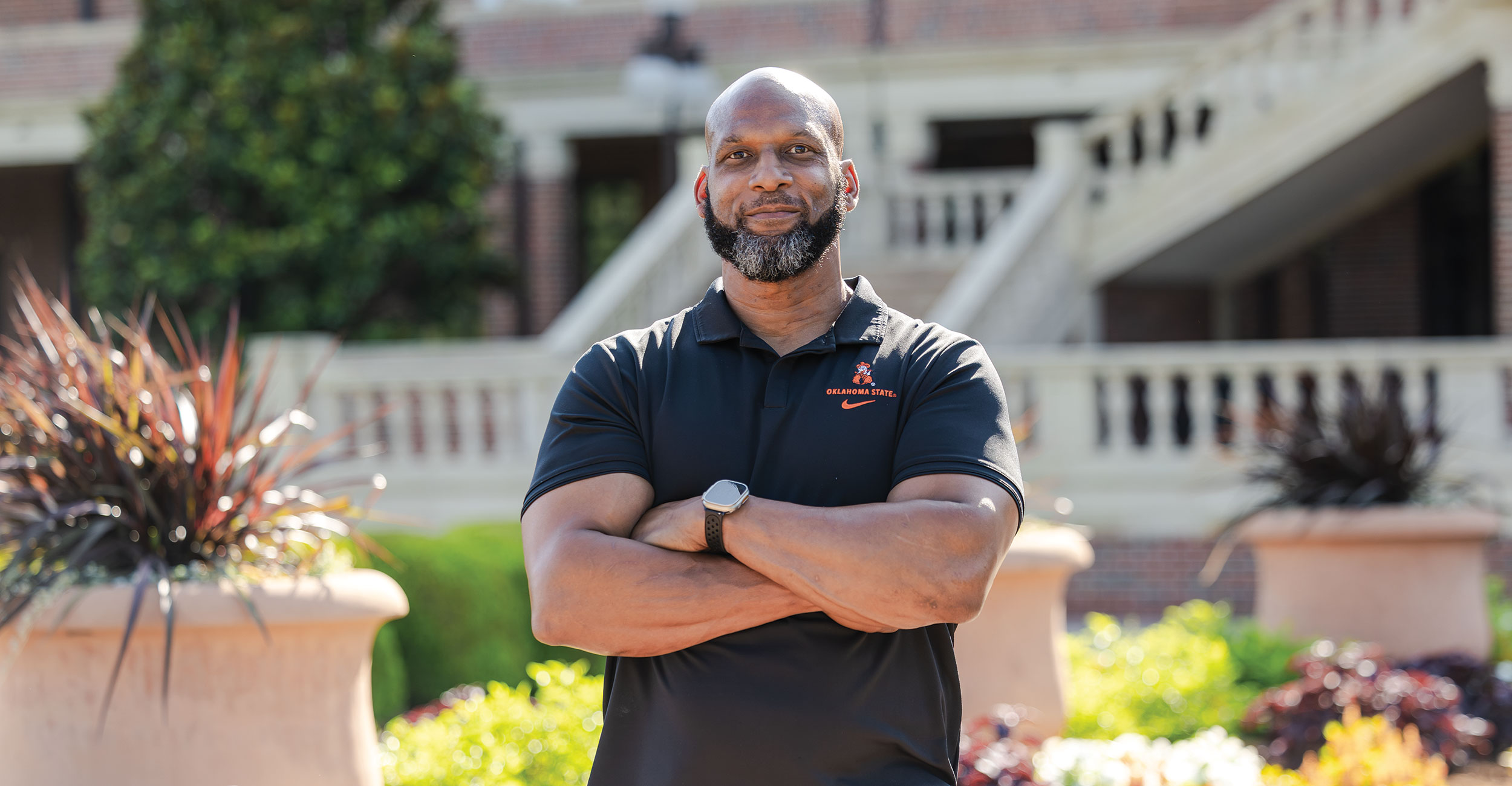 A man stands outdoors with arms crossed, wearing a black polo shirt with an orange logo. A man stands outdoors with arms crossed, wearing a black polo shirt with an orange logo.