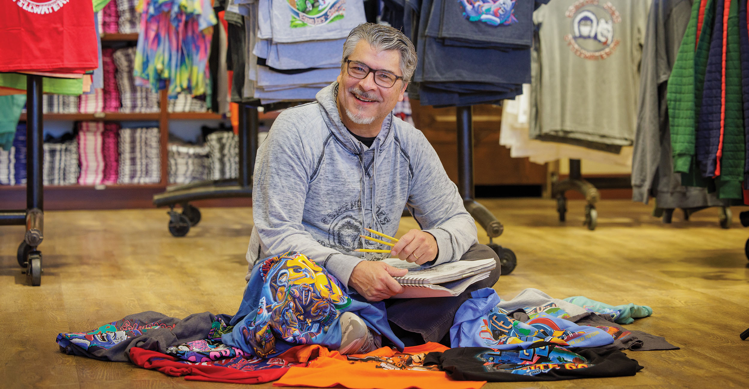A man with short gray hair, wearing a light gray hoodie and jeans, is sitting on the floor of a clothing store. He is surrounded by various colorful t-shirts laid out on the floor and is holding a notebook and pen. A man with short gray hair, wearing a light gray hoodie and jeans, is sitting on the floor of a clothing store. He is surrounded by various colorful t-shirts laid out on the floor and is holding a notebook and pen.