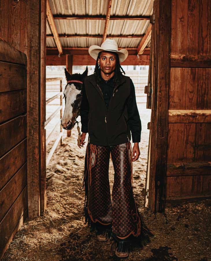 Taylor Williams stands in a wooden barn doorway wearing a cream cowboy hat, black jacket, and designer chaps with a monogram pattern, with horses visible in the stalls behind him.